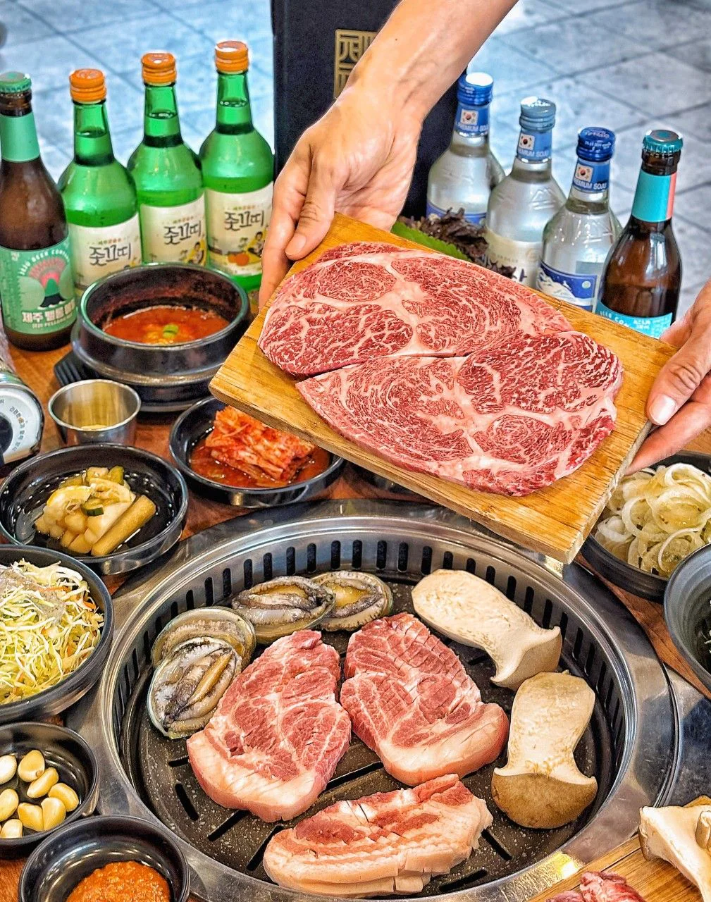 雙手端出放在木板上的韓牛,烤盤上有黑豬肉、鮑魚與菇類
